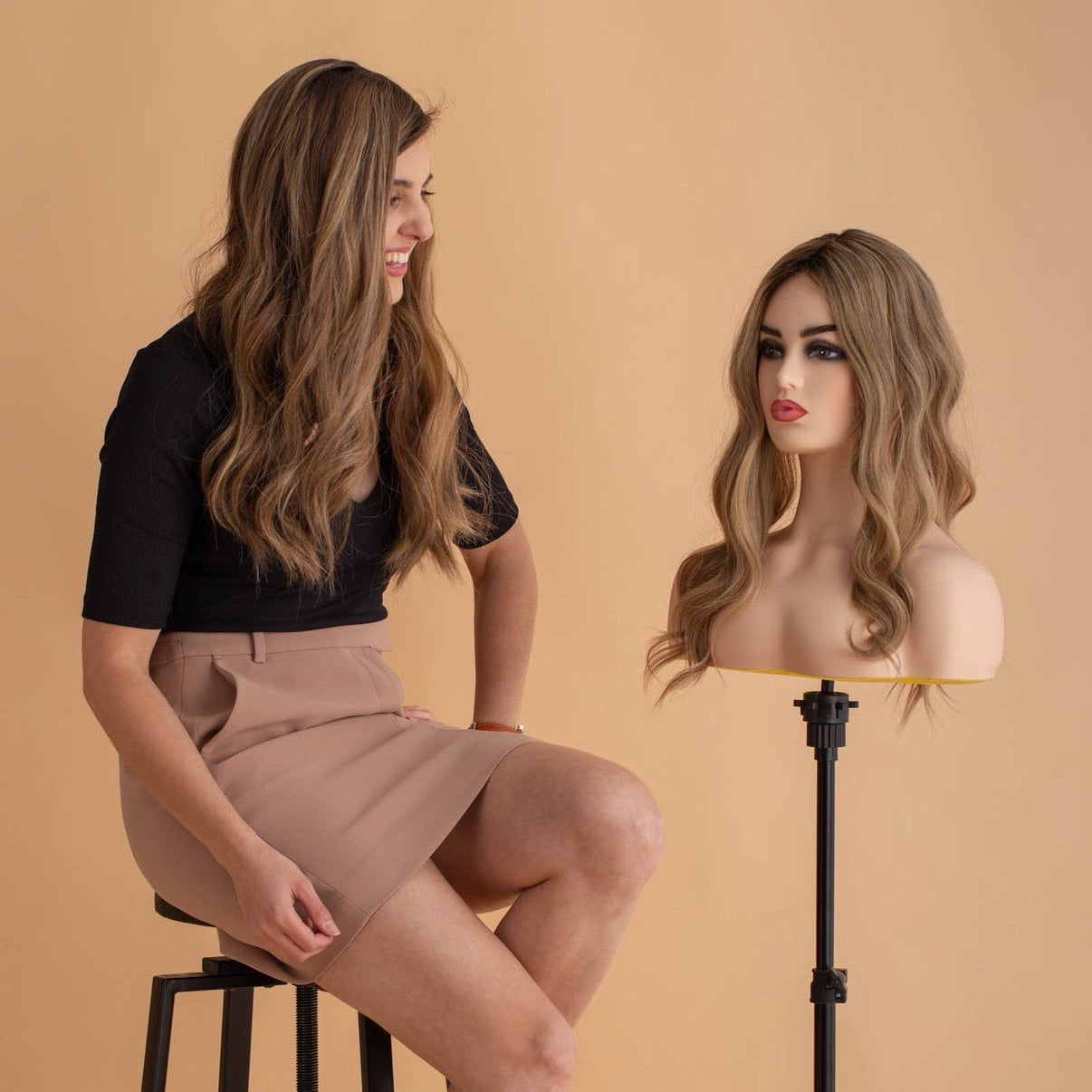 Woman sitting next to a mannequin head with a wig on a beige background