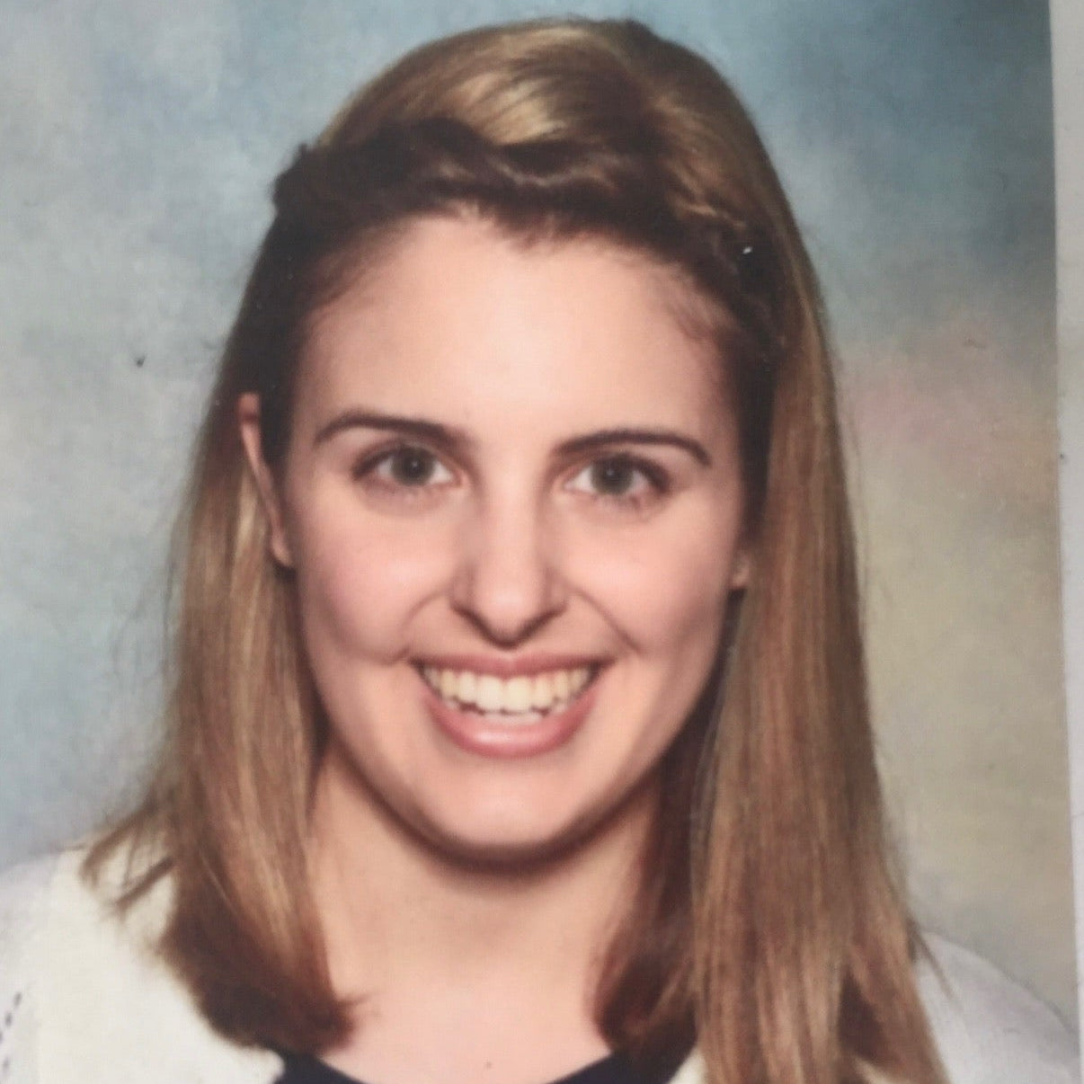 Person with shoulder-length hair wearing a white sweater over a dark top against a plain background
